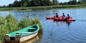 Płyną dwa kajaki po jeziorze. Stoi przycumowana łódka.