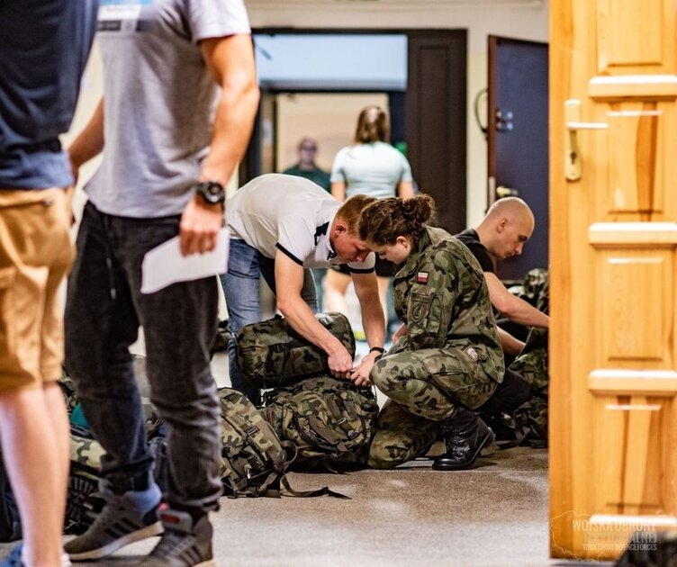 Kilka osób pakuje się do plecaka podróżnego w barwach wojskowych.