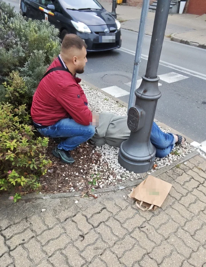 Mężczyzna w czerwonej kurtce klęczy przy leżącej osobie na pasie zieleni przy ulicy, obok stoi papierowa torba.