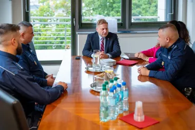 Mężczyzna w garniturze prowadzi spotkanie przy konferencyjnym stole z czterema osobami w mundurach i kobietą w różowej marynarce, w jasnym pomieszczeniu z widok