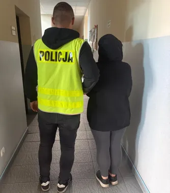 Funkcjonariusz policji w żółtej kamizelce odprowadza osobę w czarnej bluzie z kapturem korytarzem budynku.