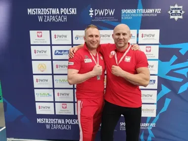 Dwaj mężczyźni w czerwonych koszulkach z medalami pozują na tle ścianki sponsorskiej podczas Mistrzostw Polski w zapasach.